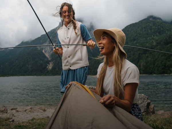 Due donne che ridono mentre montano una tenda in un paesaggio montano